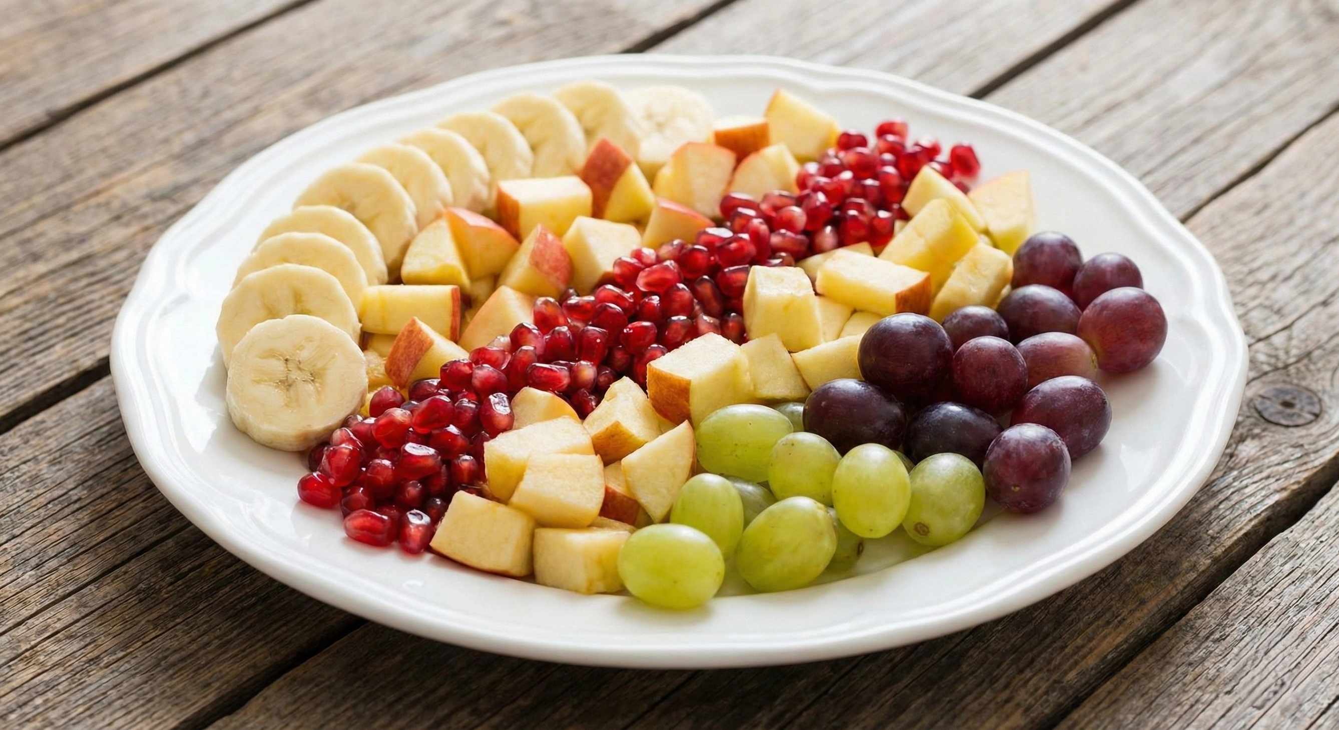 A decorative platter arranged with fresh fruit slices including bananas, apples, pomegranate seeds, and grapes for fasting energy.