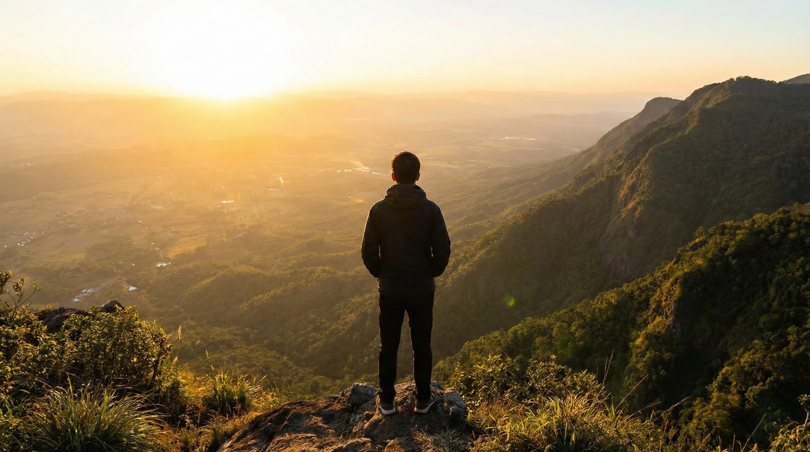 A person stands on a mountain peak at sunrise, looking out over a vast, golden valley, symbolizing a new vision and future possibilities.