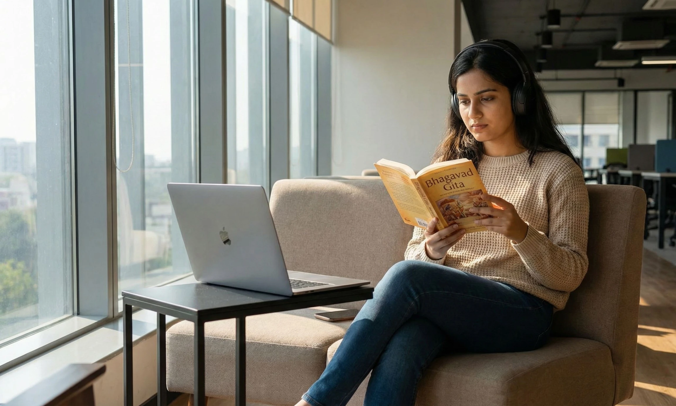 A young woman with headphones sA young woman with headphones sits by a large window in a modern office lounge, engrossed in reading a book titled 