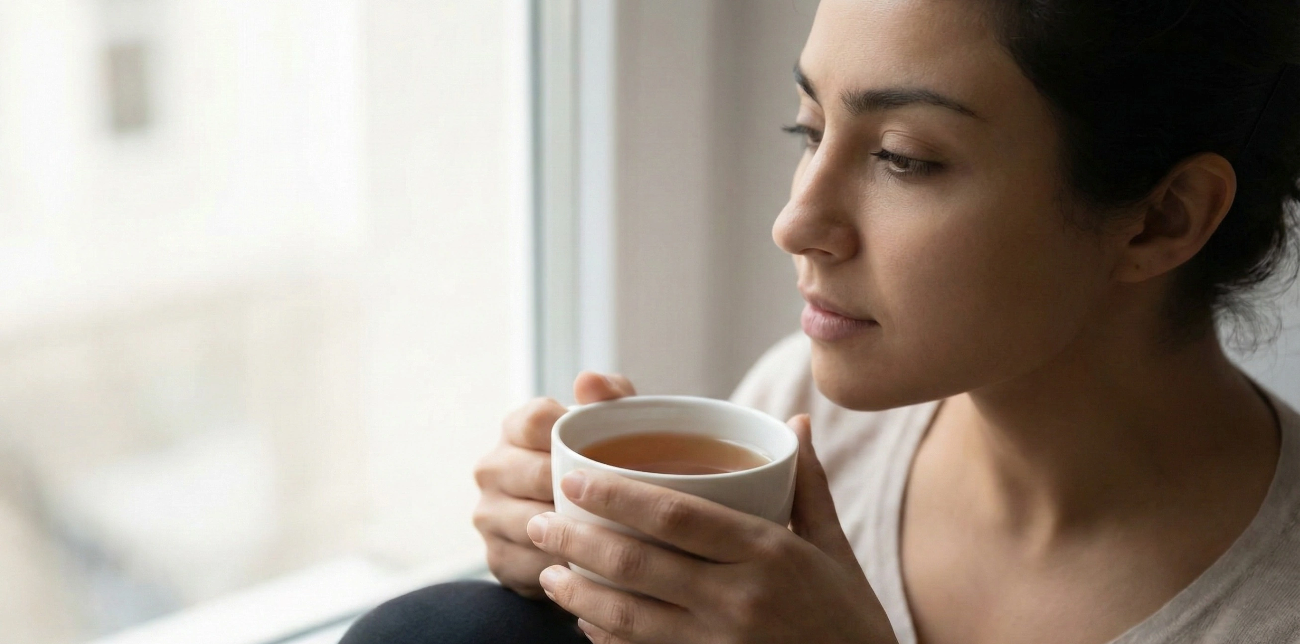 A person mindfully holding a cup of tea, looking out a window with a contemplative expression, illustrating the practice of witnessing the body