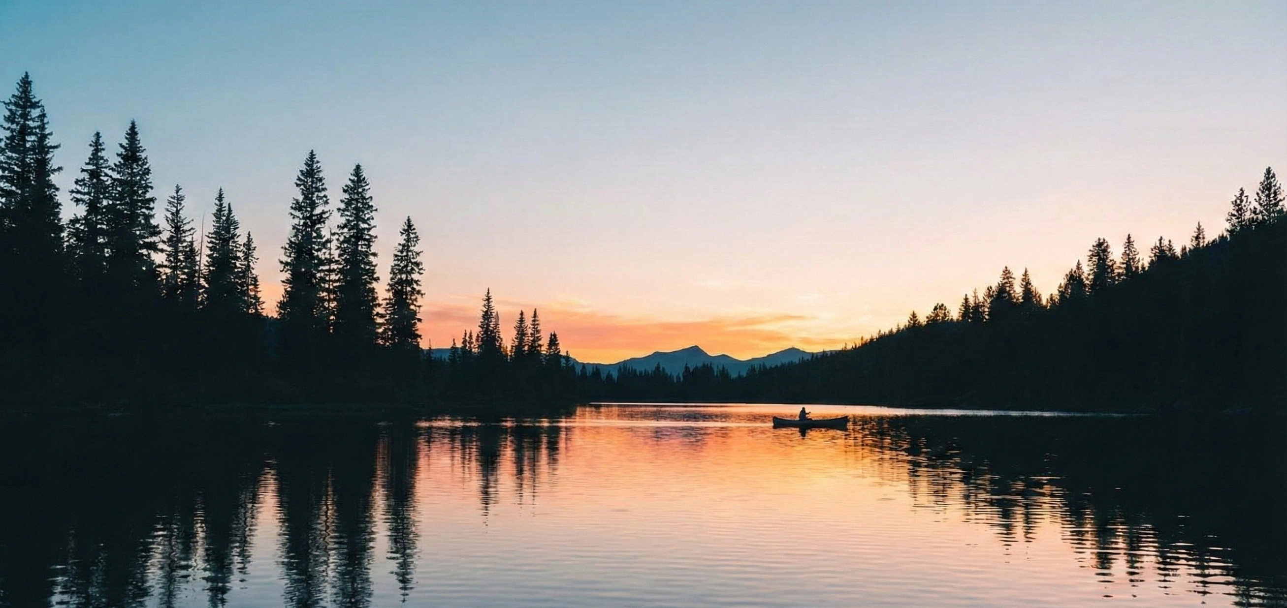 A panoramic view of a calm alpine lake at sunset, mirroring the warm orange and pink hues of the sky. Tall pine trees form dark silhouettes along the banks, and a lone canoeist drifts peacefully on the water