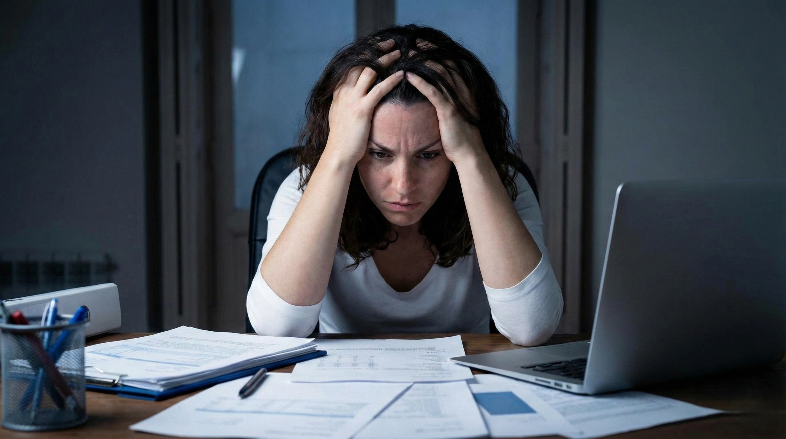 A stressed woman sitting at a cluttered desk in a dimly lit room, holding her head in her hands while looking down at piles of paperwork and a laptop, illustrating workplace burnout.