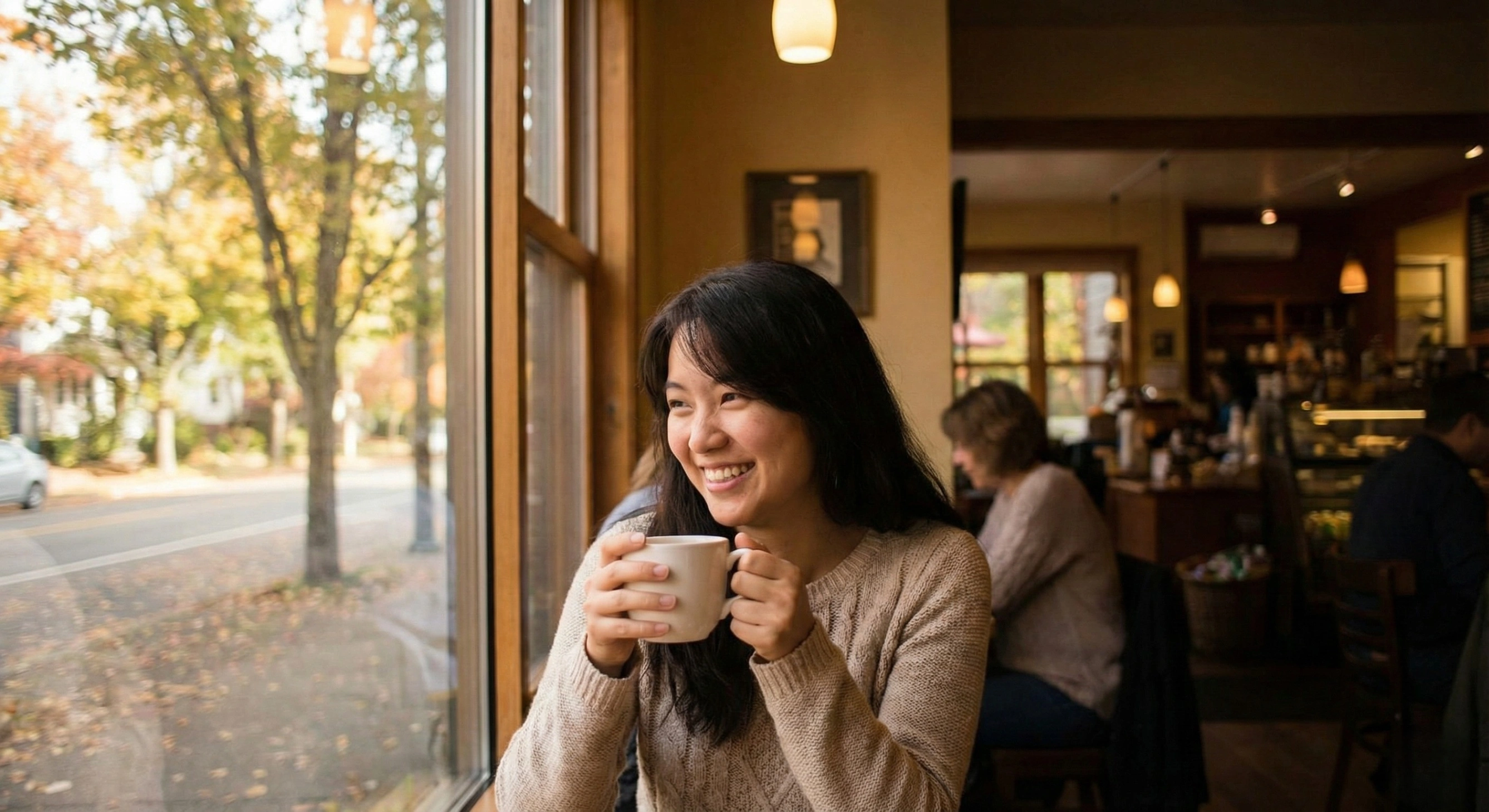 A smiling young woman holding a white mug sits in a cozy cafe next to a large window, which overlooks a street lined with golden autumn trees. The warm interior features soft lighting and wooden furniture, creating a peaceful, seasonal atmosphere.