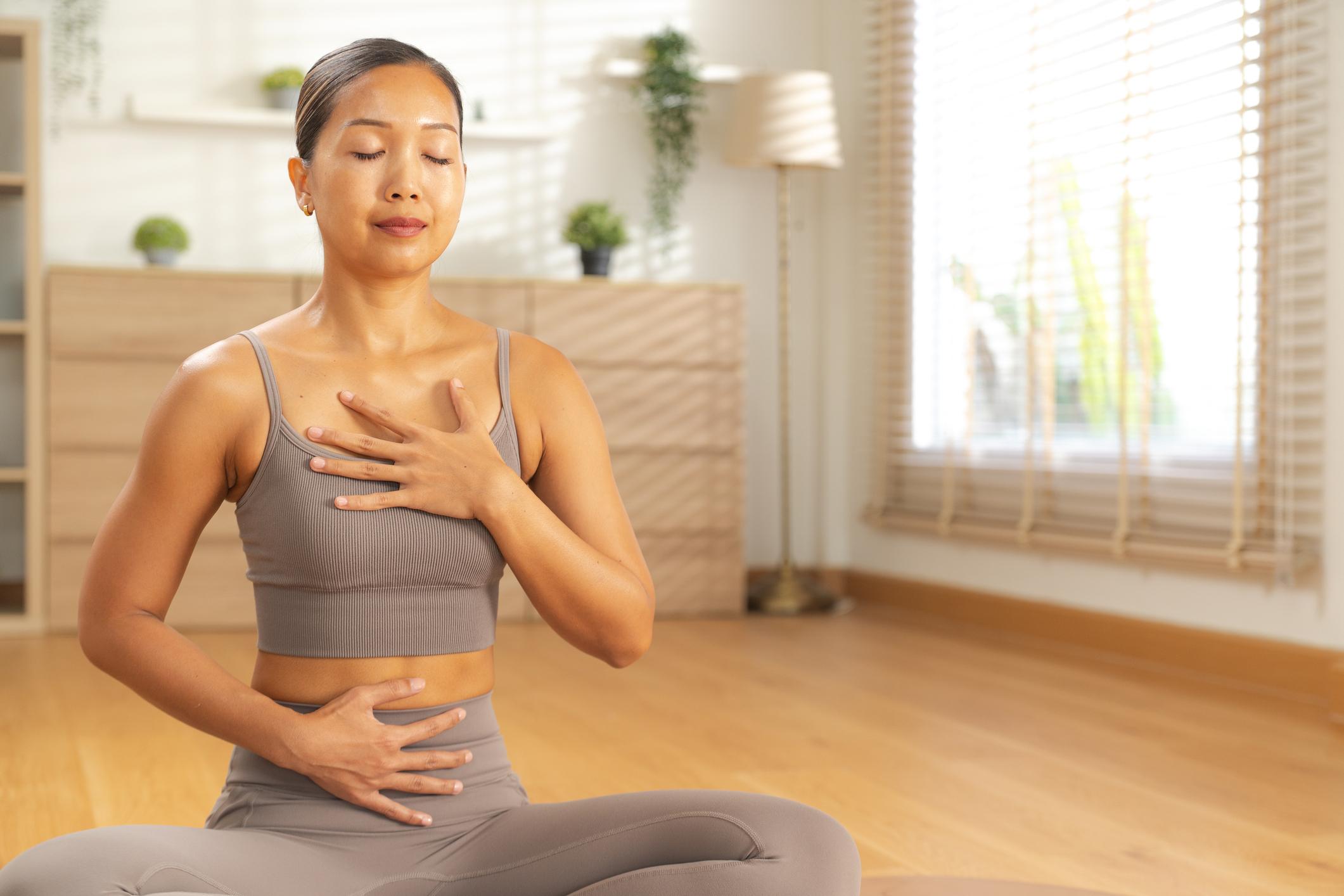 Young woman practicing mindful breathing in sunlit living room, hand on chest and abdomen, calm meditation with natural light